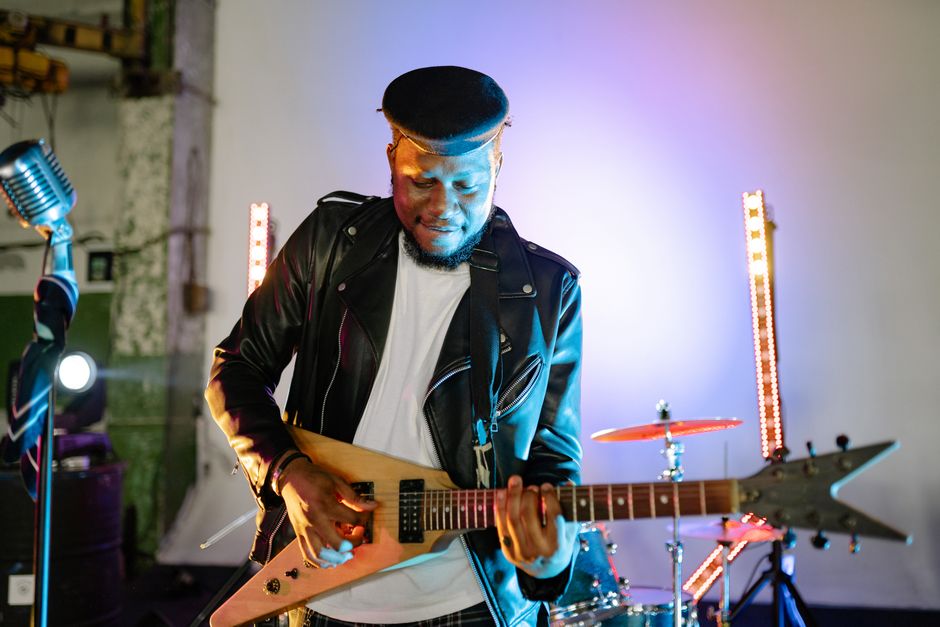 Guitarist playing electric guitar on stage with vibrant lights, wearing a leather jacket and flat cap.