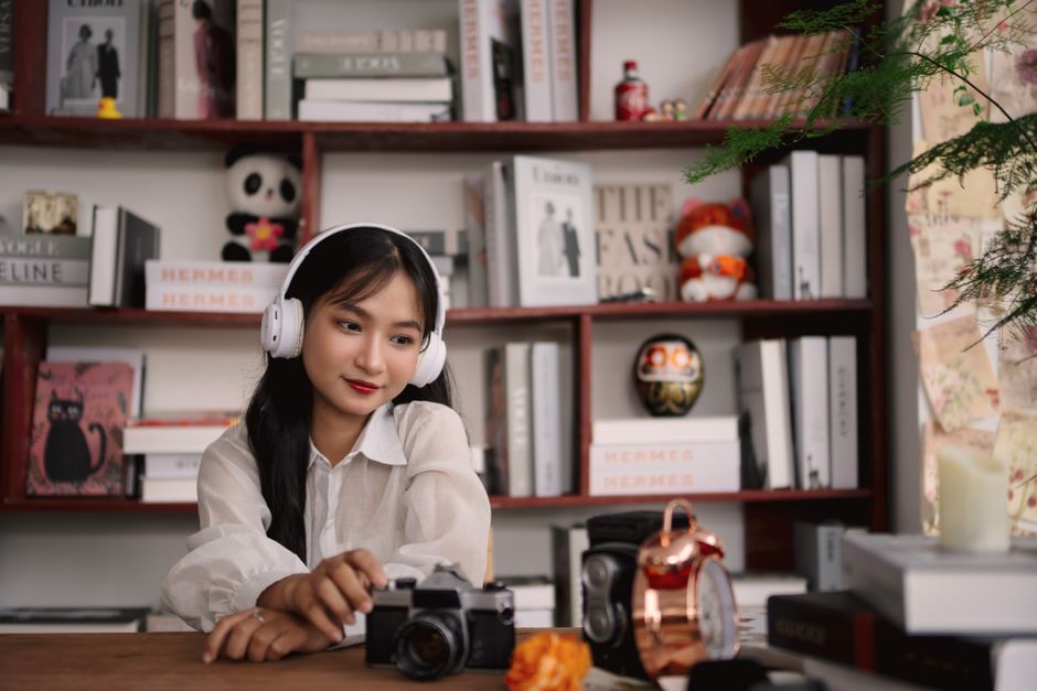 Asian woman enjoying music, sitting indoors with a camera in a cozy book-filled room.
