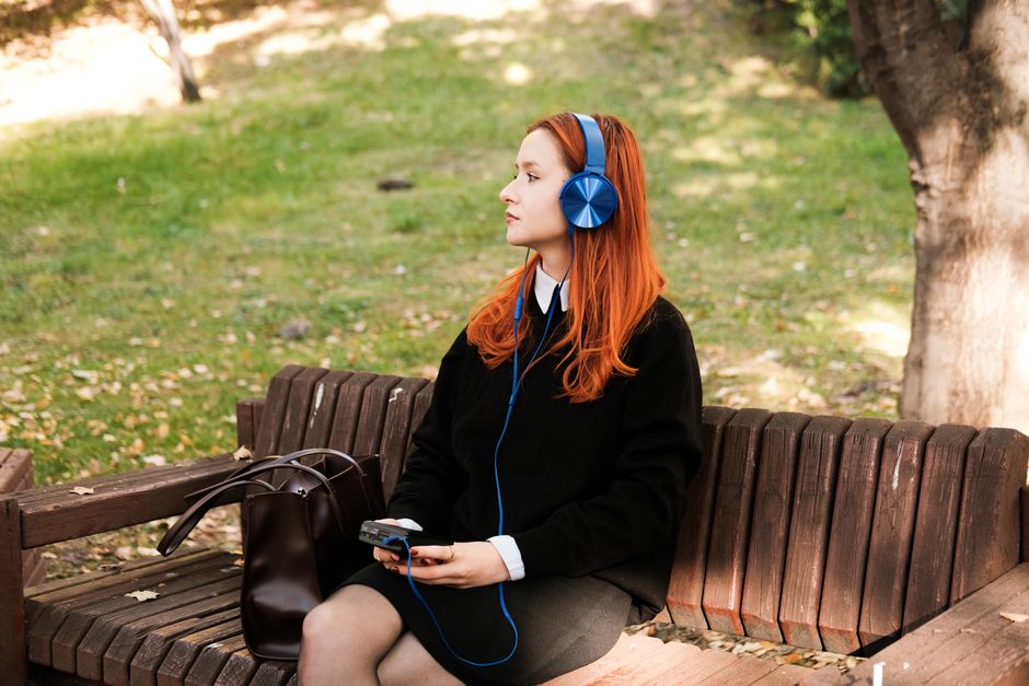 A young woman with red hair wearing headphones sits on a bench in a sunny park.