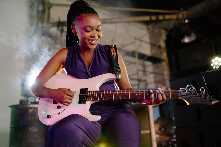 Joyful woman musician plays electric guitar with a smile in a dimly lit indoor setting, showcasing talent and passion.