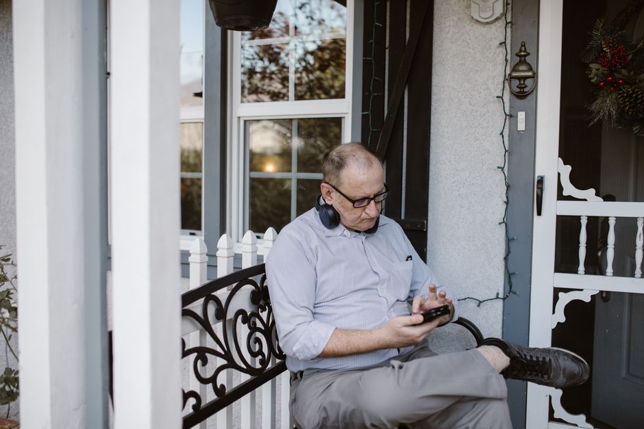 Elderly man with headphones browsing phone while sitting on a porch bench.