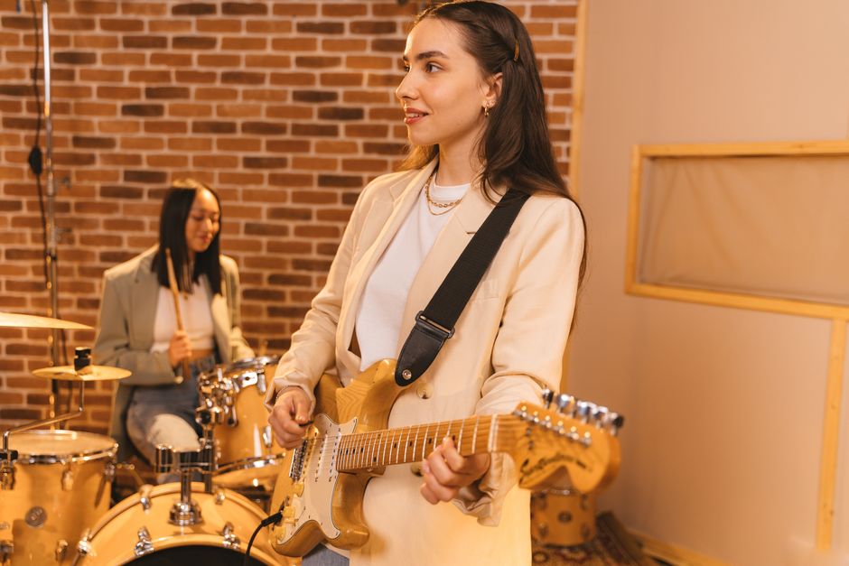 Two women musicians playing guitar and drums indoors with a brick wall backdrop.