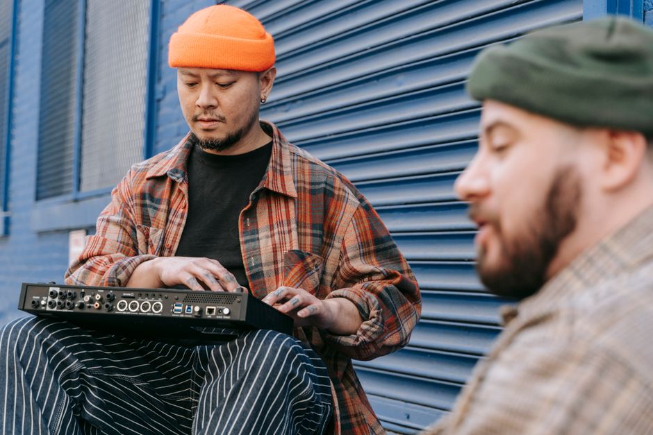 Two musicians with a synthesizer outside, one wearing an orange hat, in front of a blue garage door.