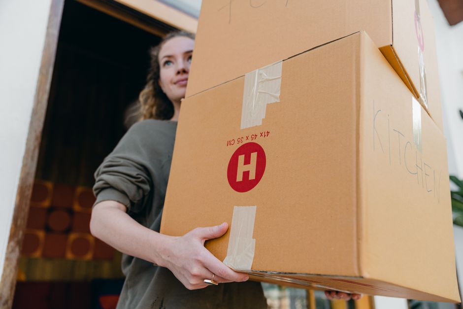 From below of young slim woman carrying stack of heavy carton boxes with kitchen stuff and looking away while moving out of old house