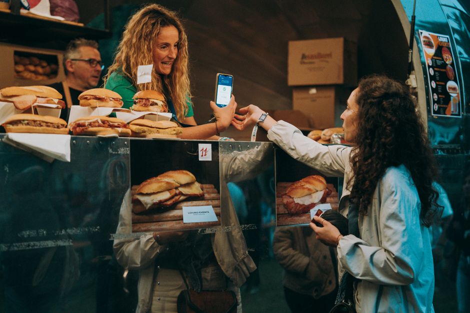 Two women exchanging digital payment at a food truck selling sandwiches outdoors in a bustling setting.