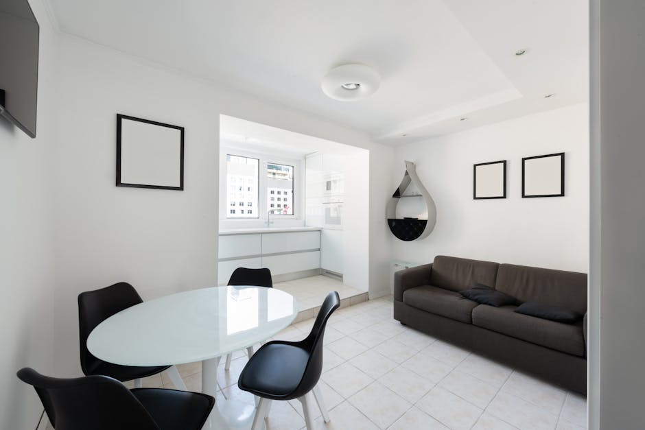 Interior of modern light house with glass table near chairs and couch with pillows near paintings and shelves on wall near entrance to kitchen with counter with window
