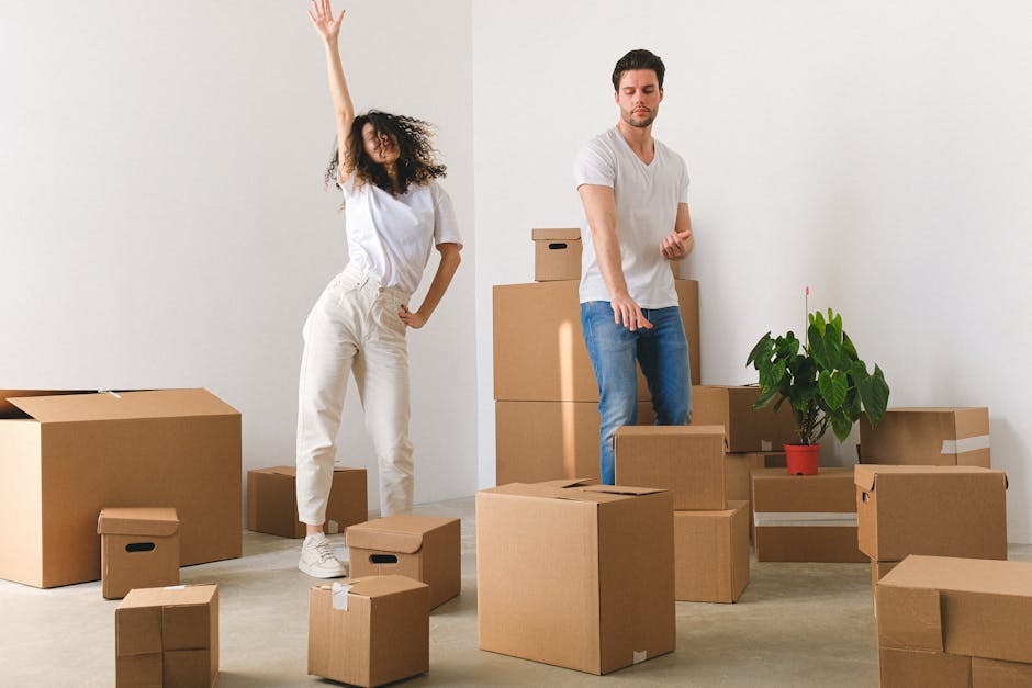 Full length of cheerful young man and woman in casual clothes dancing happily amidst cardboard boxes after relocation into new light apartment