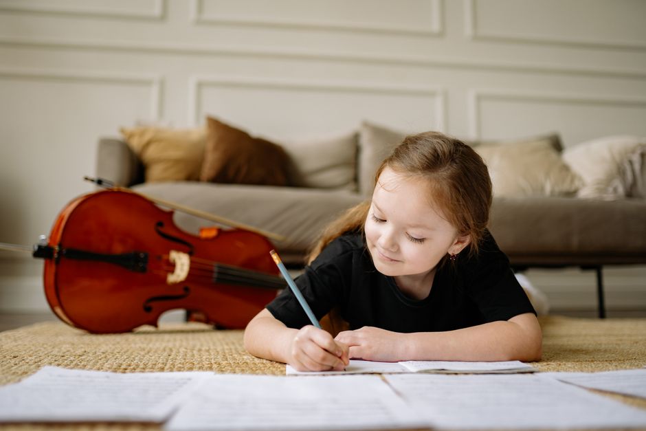A young girl lies on the floor writing in a notebook, with a cello in the background, creating a cozy indoor setting.