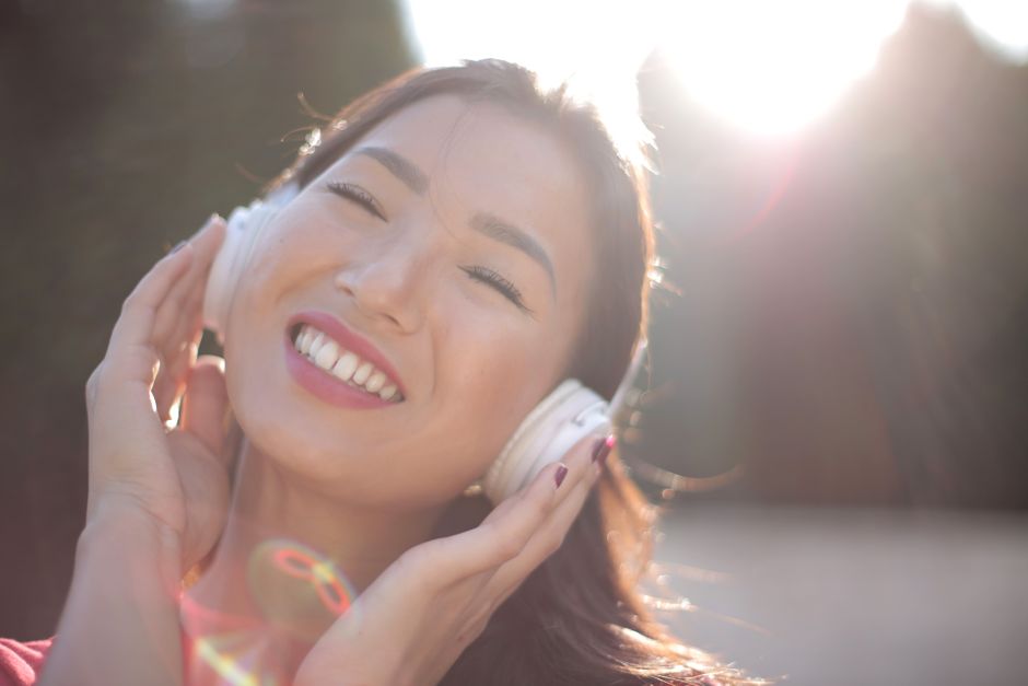 Bright portrait of an Asian woman enjoying music outdoors with headphones, radiating happiness.