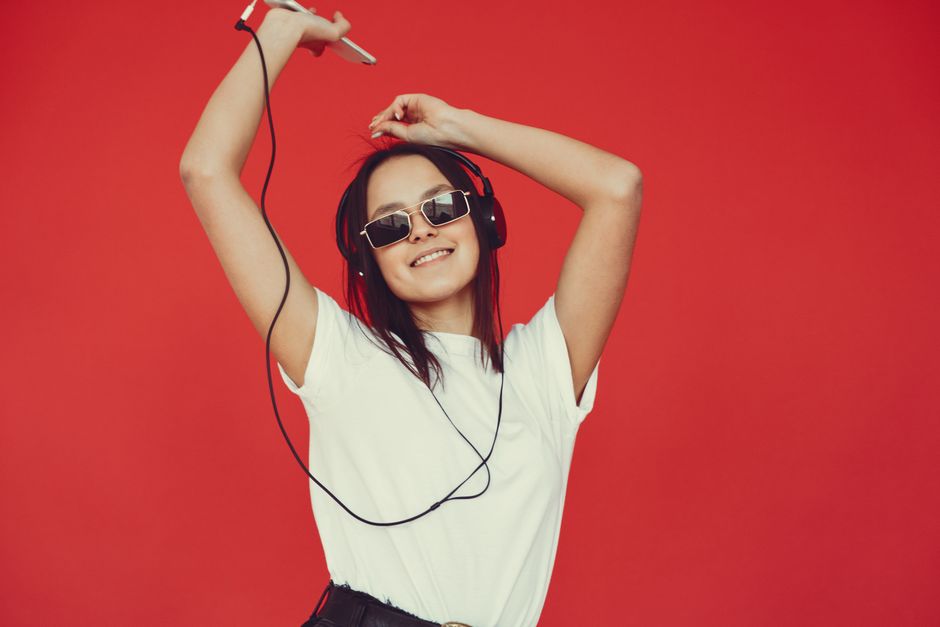Optimistic positive millennial female in white top and stylish sunglasses listening to favorite playlist and looking at camera while dancing with raised arms in studio on red background