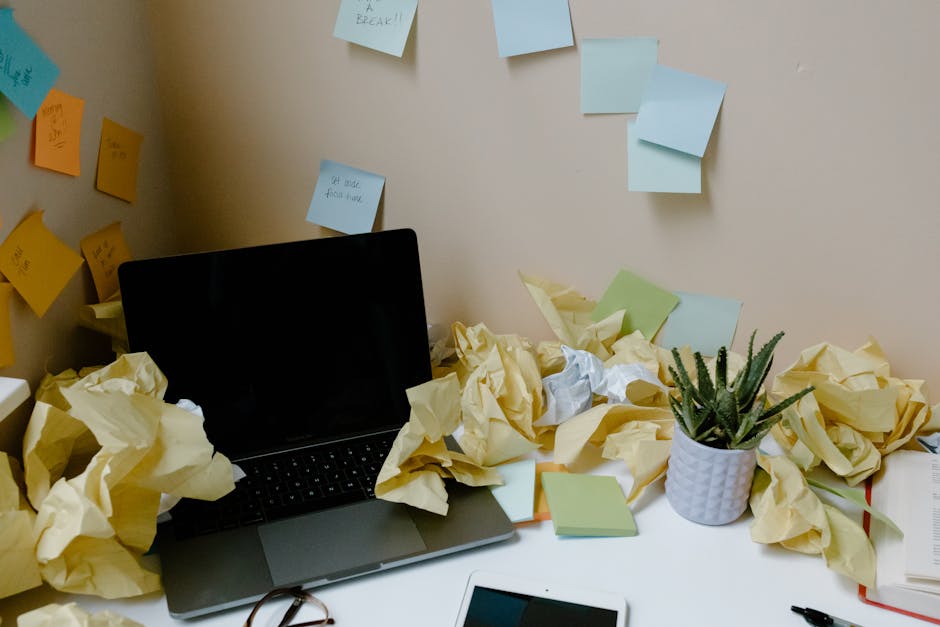 A chaotic office desk with a laptop, scattered papers, sticky notes, and a potted plant.