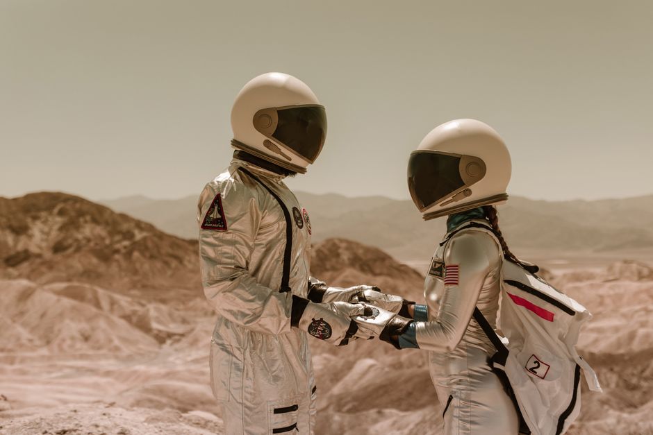 Two astronauts in full spacesuits holding hands in a barren, desert-like landscape symbolizing space exploration.