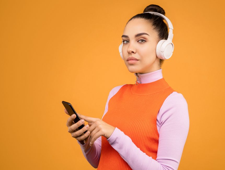 Portrait of a stylish woman using a smartphone and wearing headphones against an orange background.